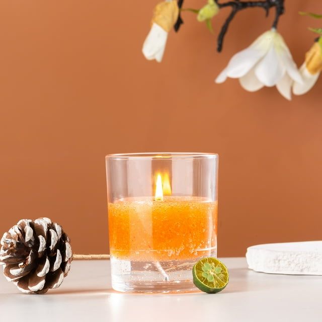 Lit candle in a glass container with orange wax, surrounded by decorative elements like a pine cone and lime on a brown background