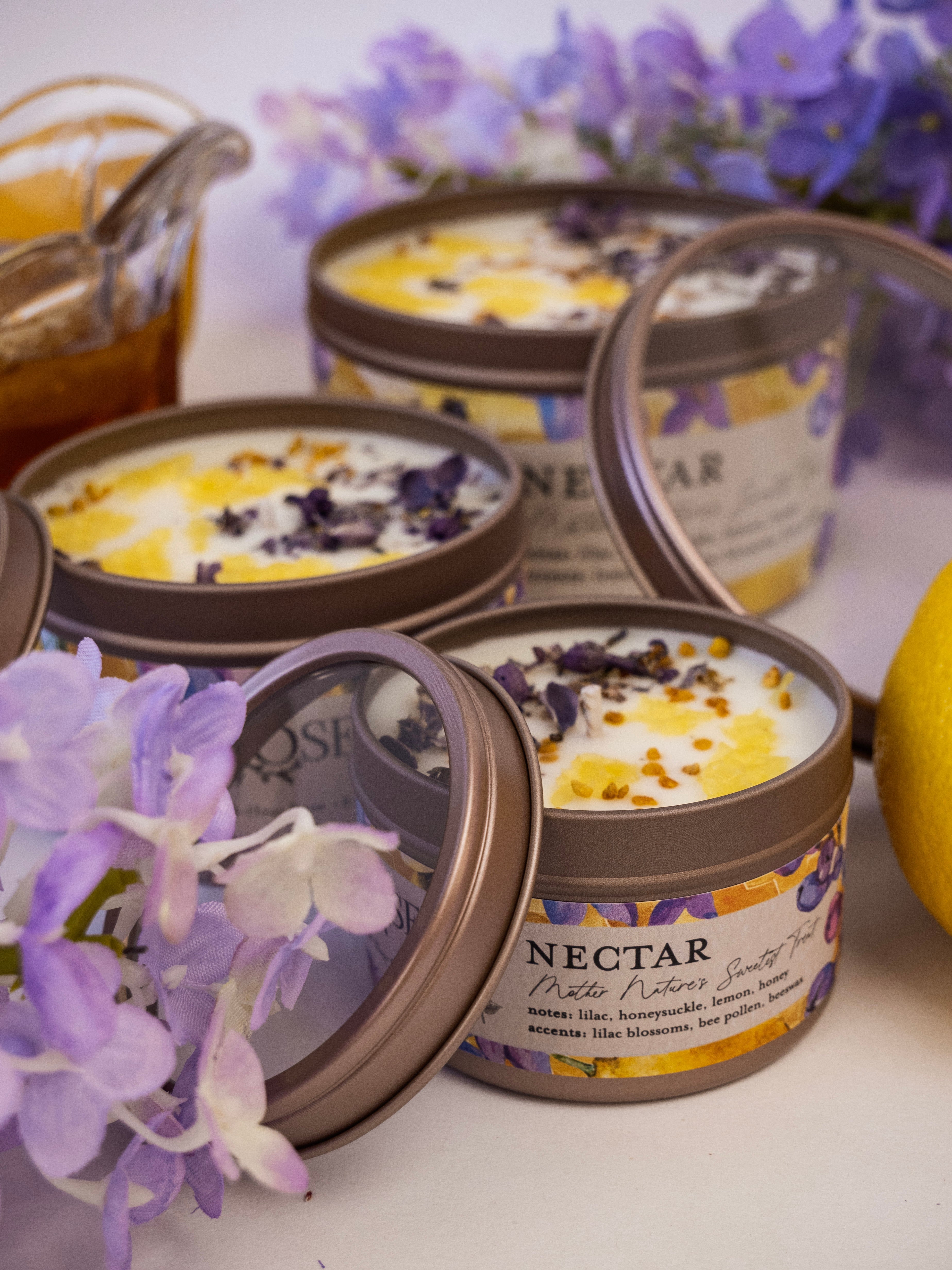 NECTAR candles in tins with labels and lids off surrounded by purple flowers and lemon on white background