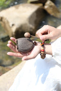 Hand holding small wooden mallet and carved fruit shell percussion instrument with colorful beaded wrist strap, rocks and water in background - natural hand percussion woodblock