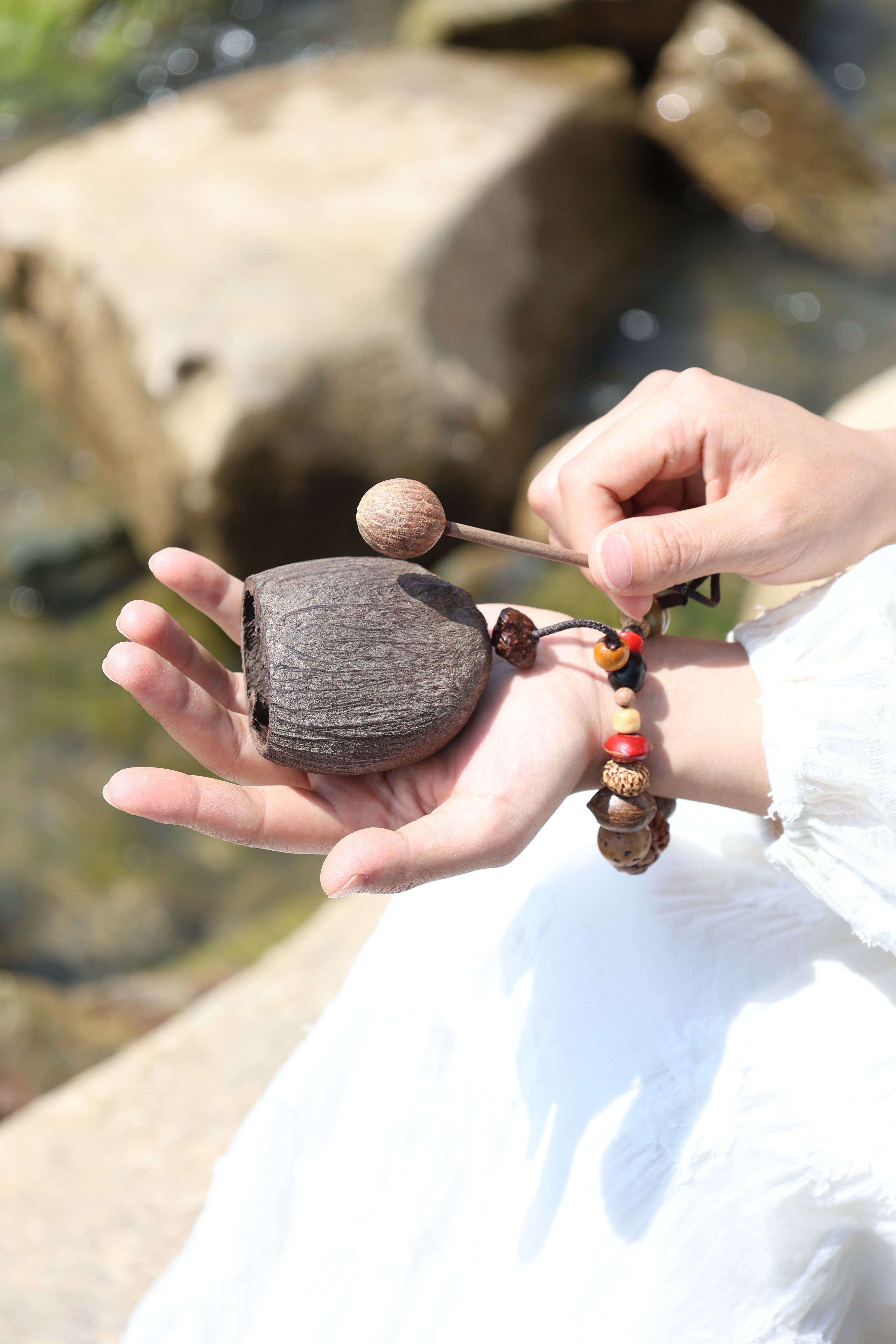 Hand holding small wooden mallet and carved fruit shell percussion instrument with colorful beaded wrist strap, rocks and water in background - natural hand percussion woodblock