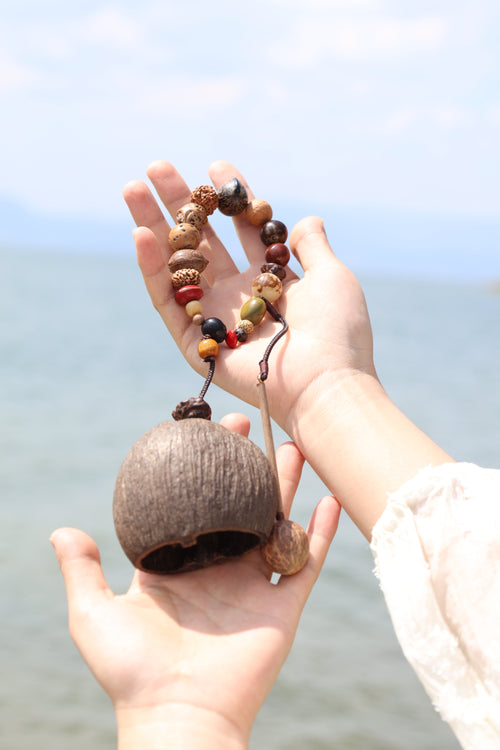 Hands holding hollowed out fruit shell percussion instrument and colorful beaded wrist strap with blurred background - handmade natural woodblock instrument