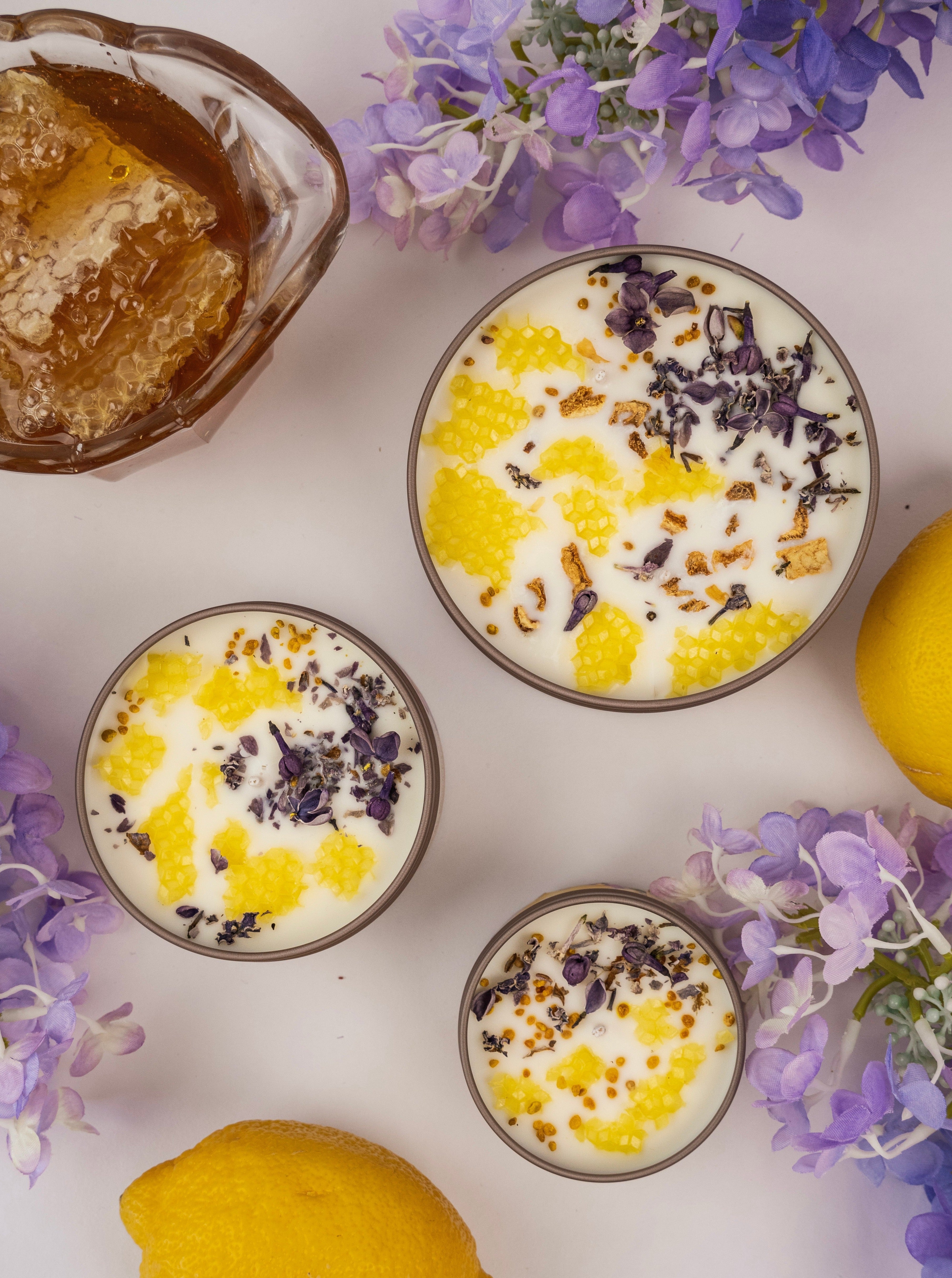 Three different sized NECTAR candles in metal tins with honeycomb, lavender, and lemon surrounded by honeycomb, lavender sprigs, and whole lemons - top view