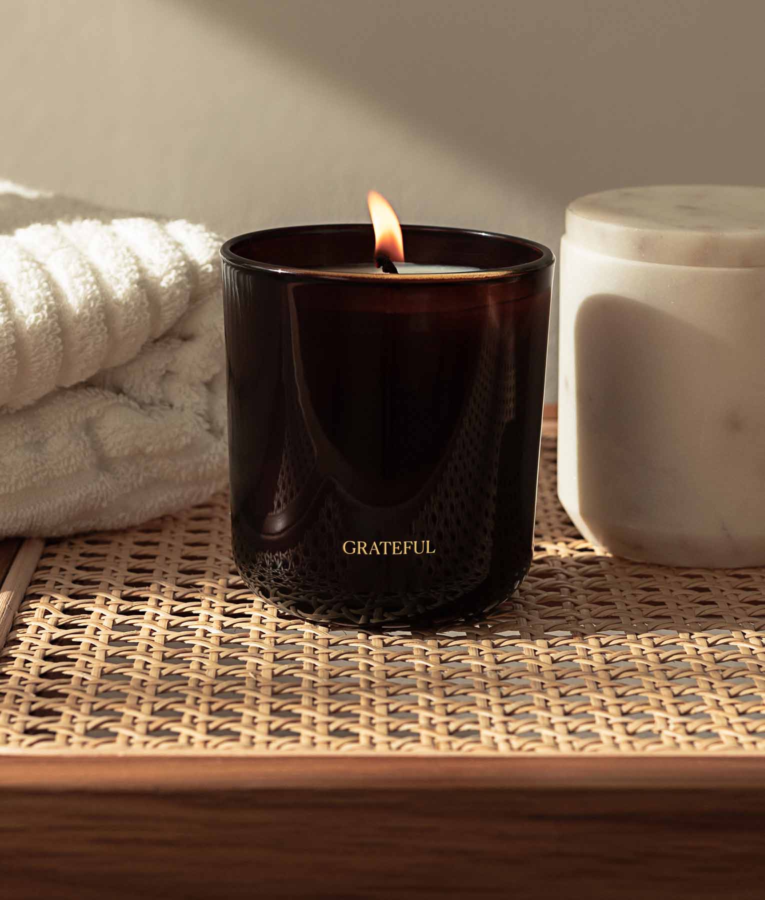 Lit Grateful candle on woven surface with towel and container in background - aromatherapy soy candle with spearmint lemongrass lavender cedarwood