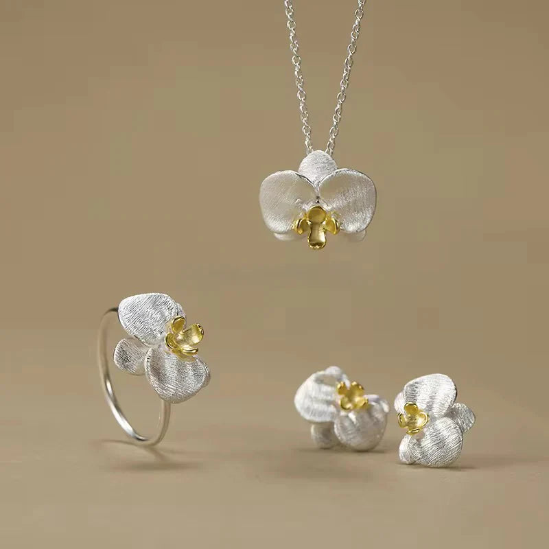 Silver orchid jewelry set with gold center accents - necklace, ring, and stud earrings on beige background
