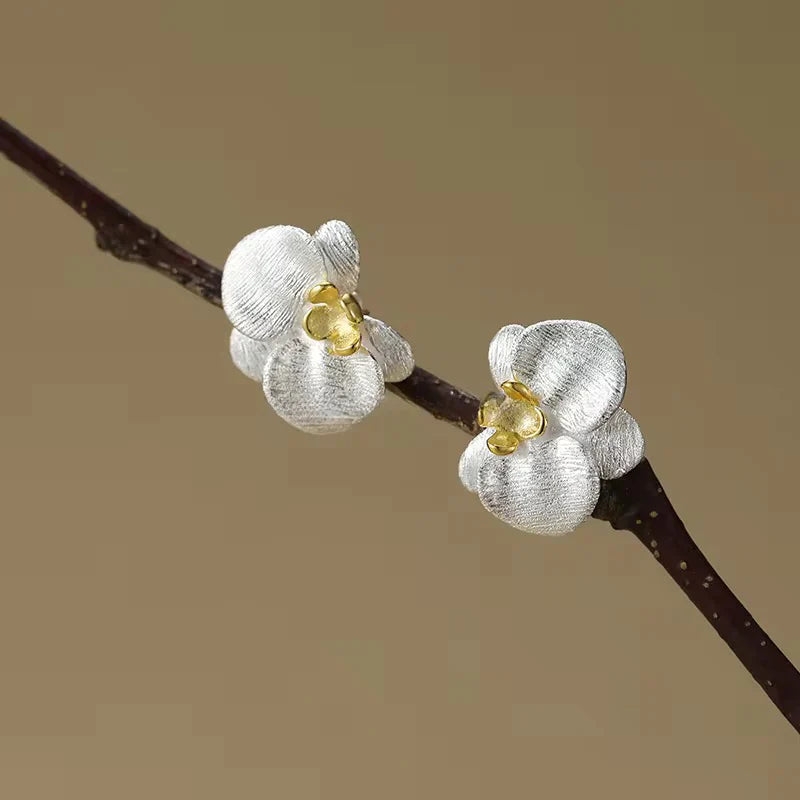Silver orchid stud earrings with gold centers on branch against brown background - 925 sterling silver