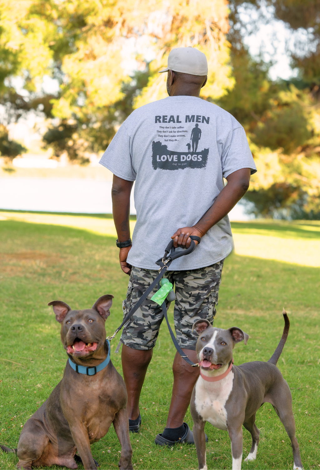 Back view of man wearing 'Real Men Love Dogs' t-shirt standing in park with two dogs on leash - dog lover graphic tee lifestyle shot