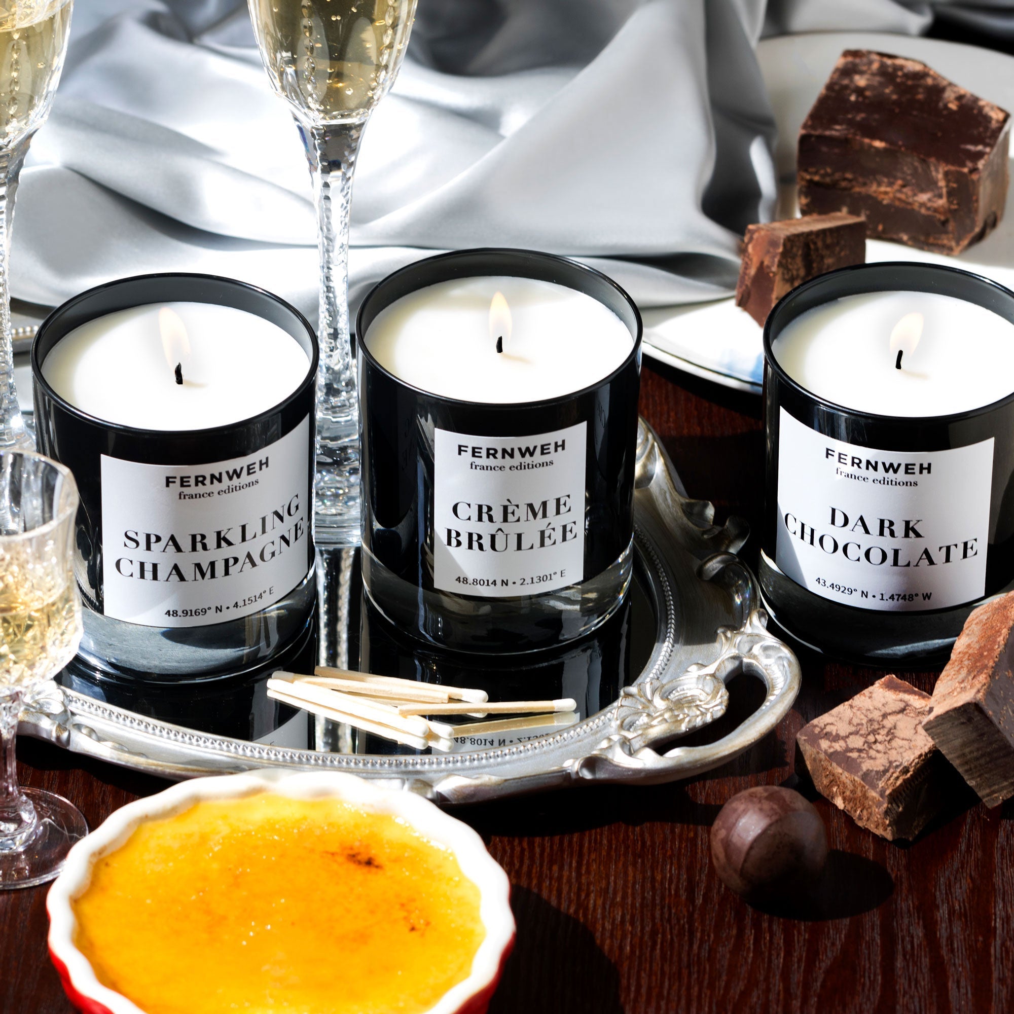 Three lit Fernweh candles - Sparkling Champagne, Crème Brûlée, and Dark Chocolate - on tray with glasses and dessert
