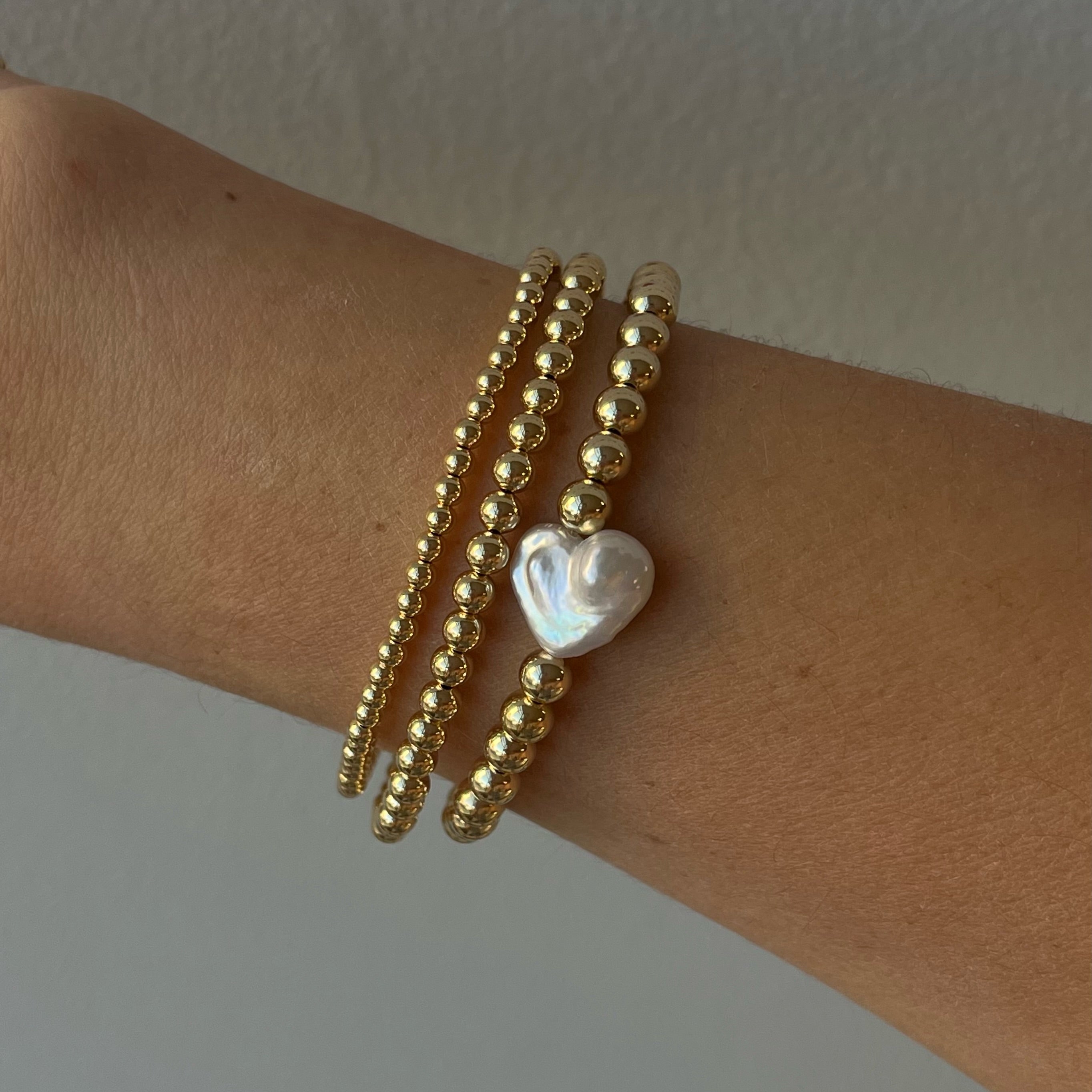 Gold beaded bracelet with heart-shaped pearl charm on wrist against neutral background showing Valentine's Day jewelry stretch design