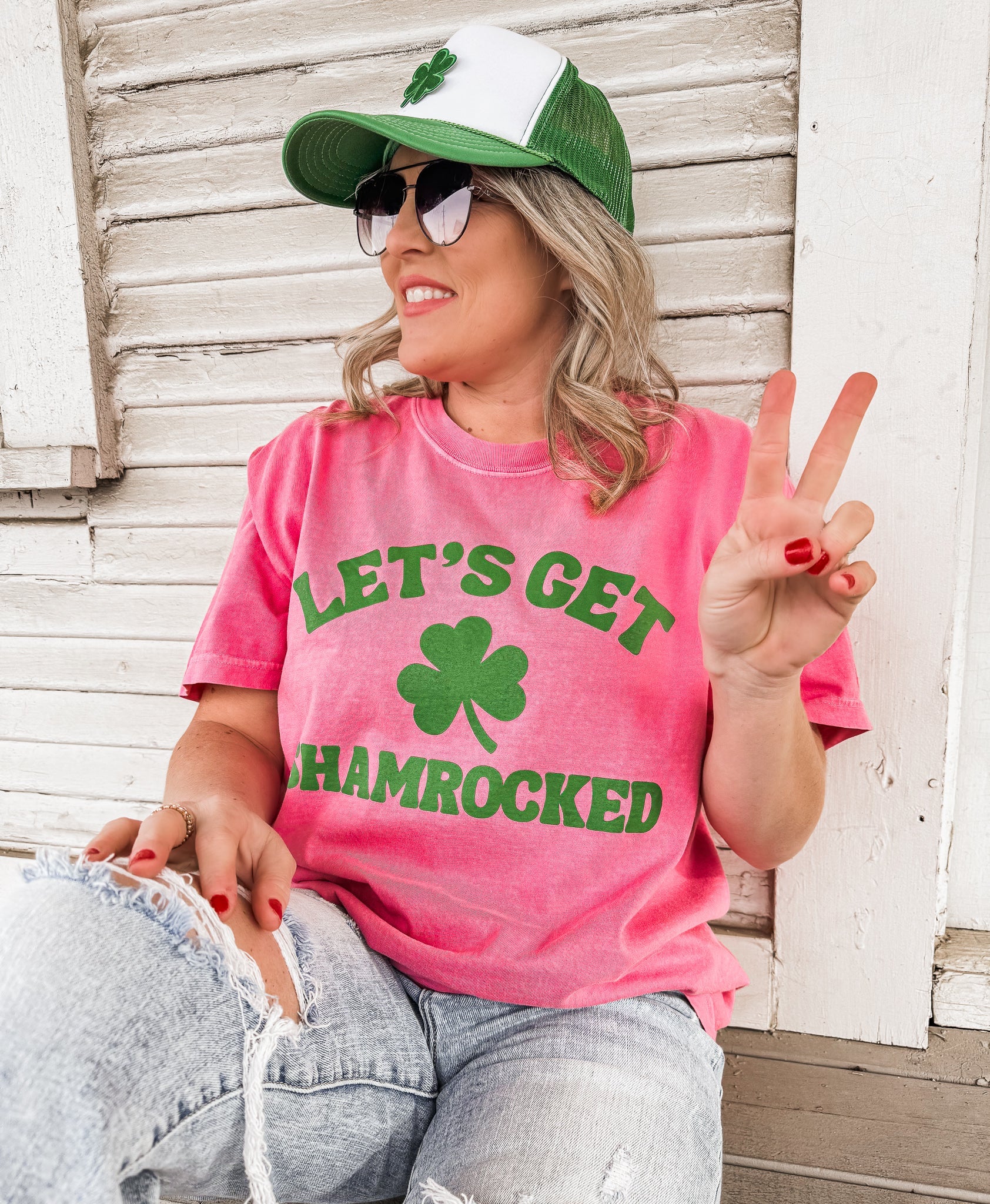 Person wearing a Crunchberry Pink t-shirt with green 'Let's Get Shamrocked' text and a green cap, sitting against a wooden wall.