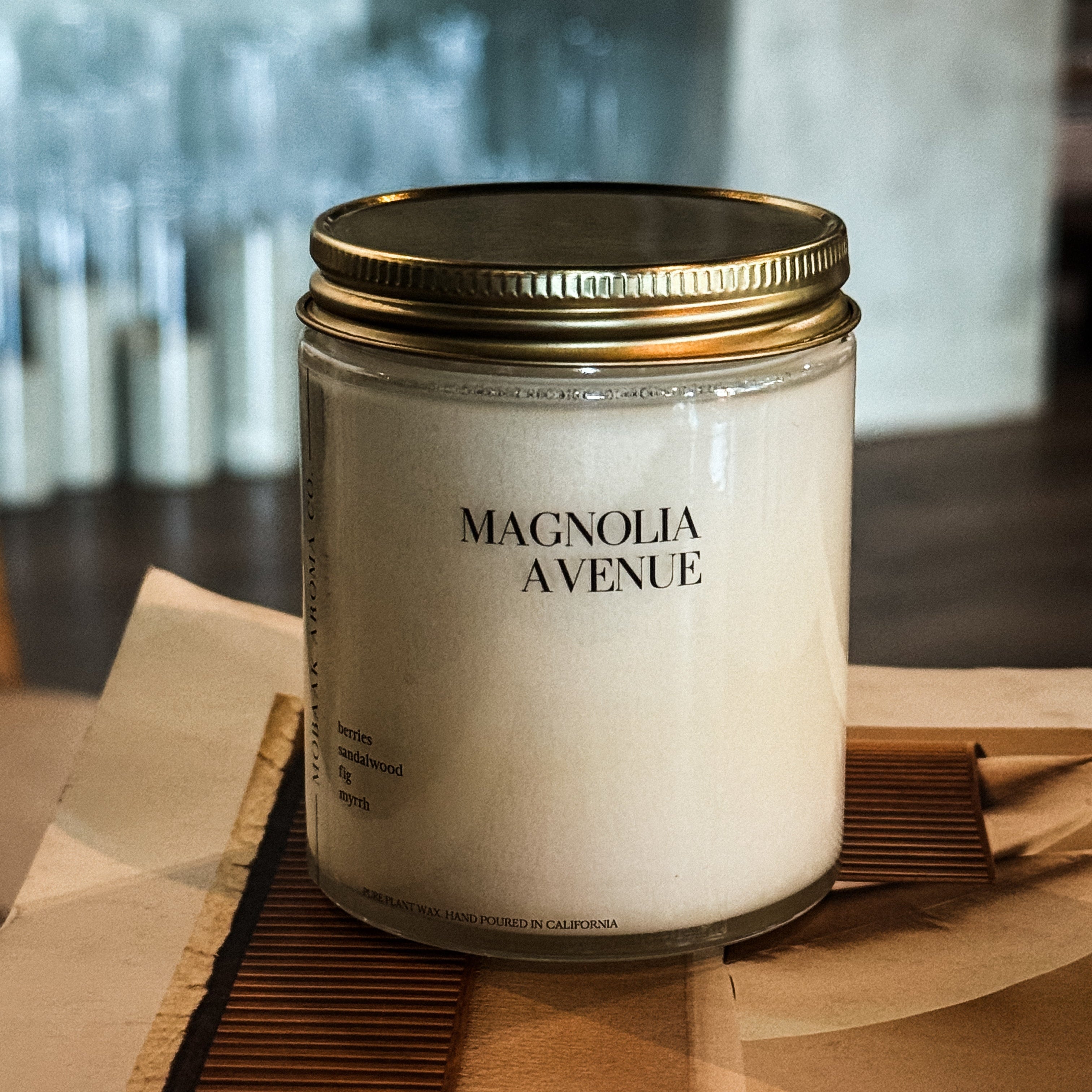Magnolia Avenue soy candle in glass jar with gold lid on wooden surface with blurred background - front view