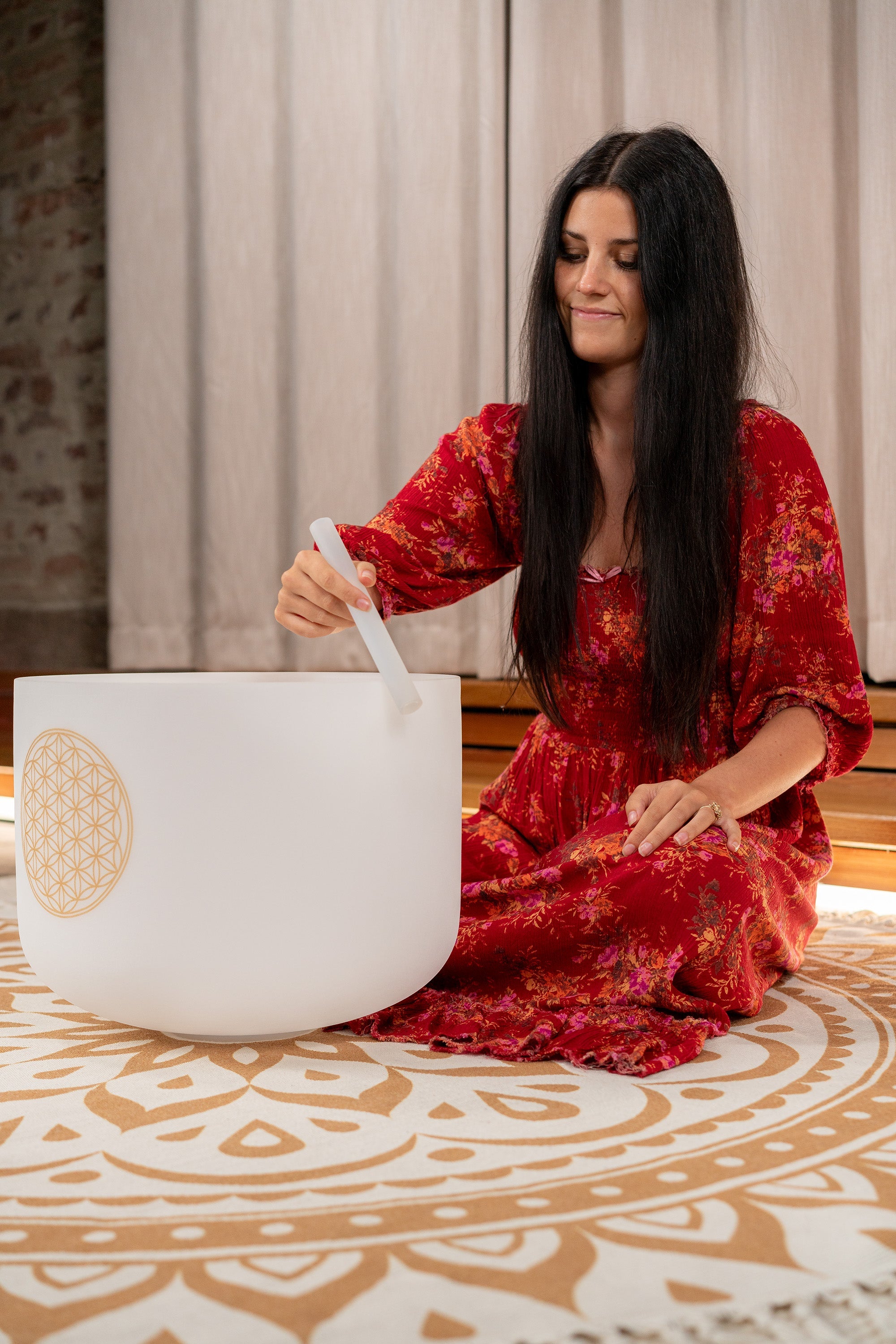 Woman in red dress playing white frosted crystal singing bowl during meditation practice in room with curtains