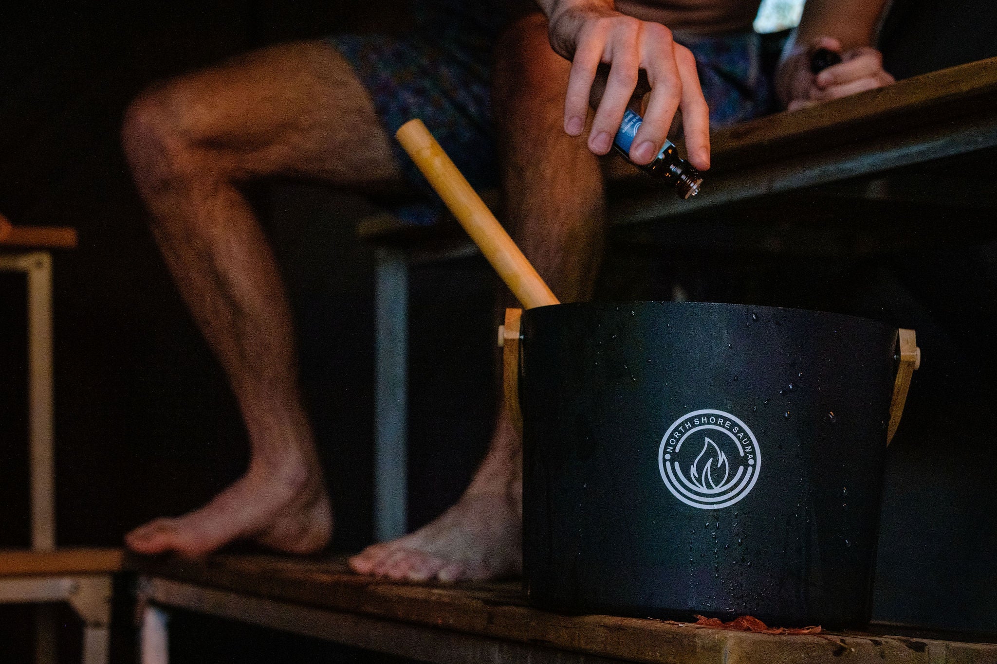 Close up view of man sitting on a wooden bench with a black pot in front of them, holding an essential oil bottle placing a drop in it, in a sauna setting