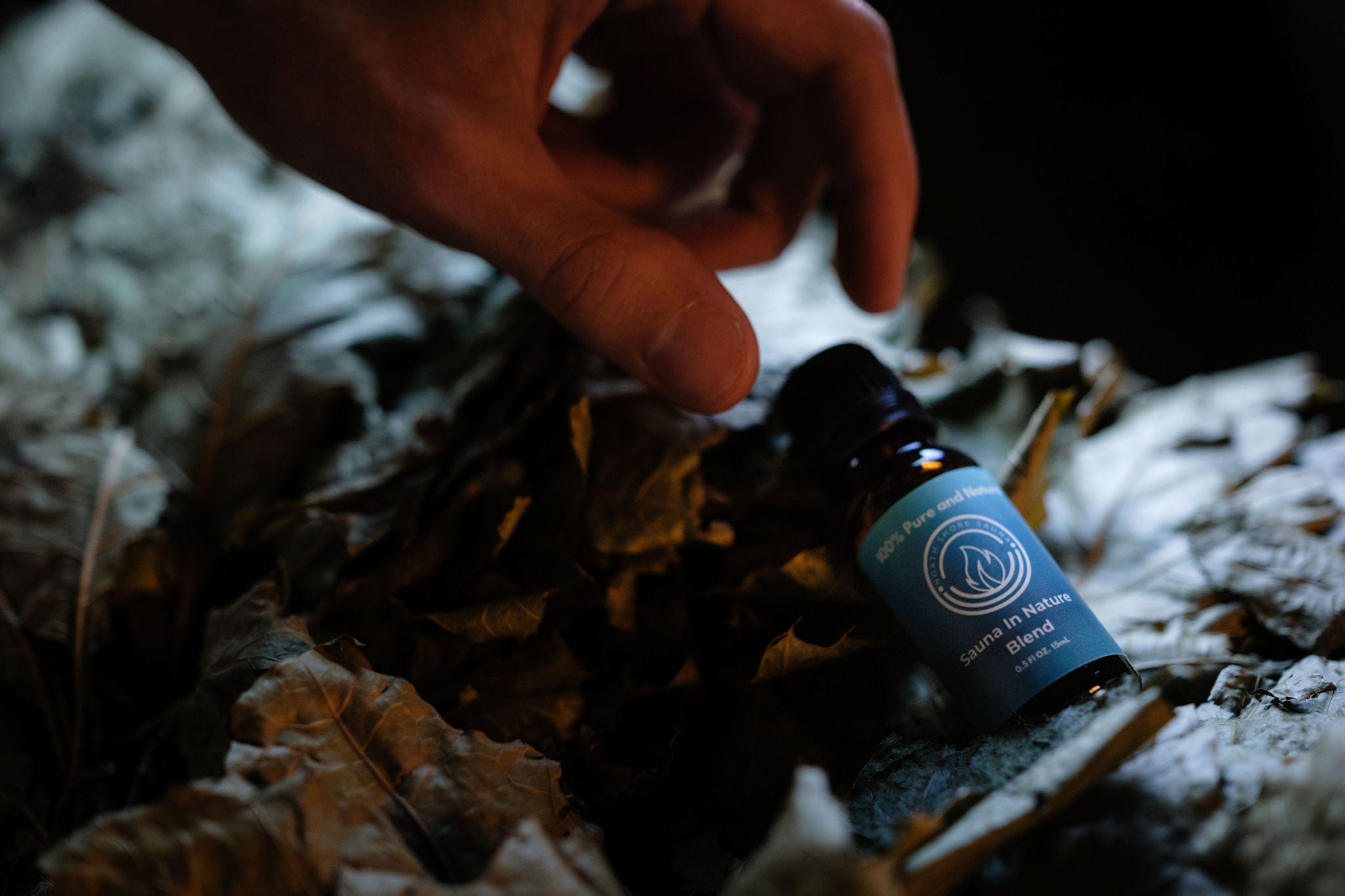 Bottle of Sauna in Nature essential oil with blue label on a leafy surface with a hand reaching towards it