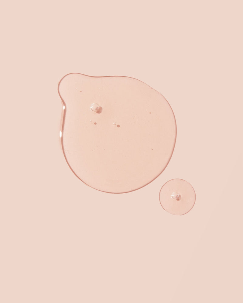 Two drops of clear liquid organic castile soap base on a light pink surface