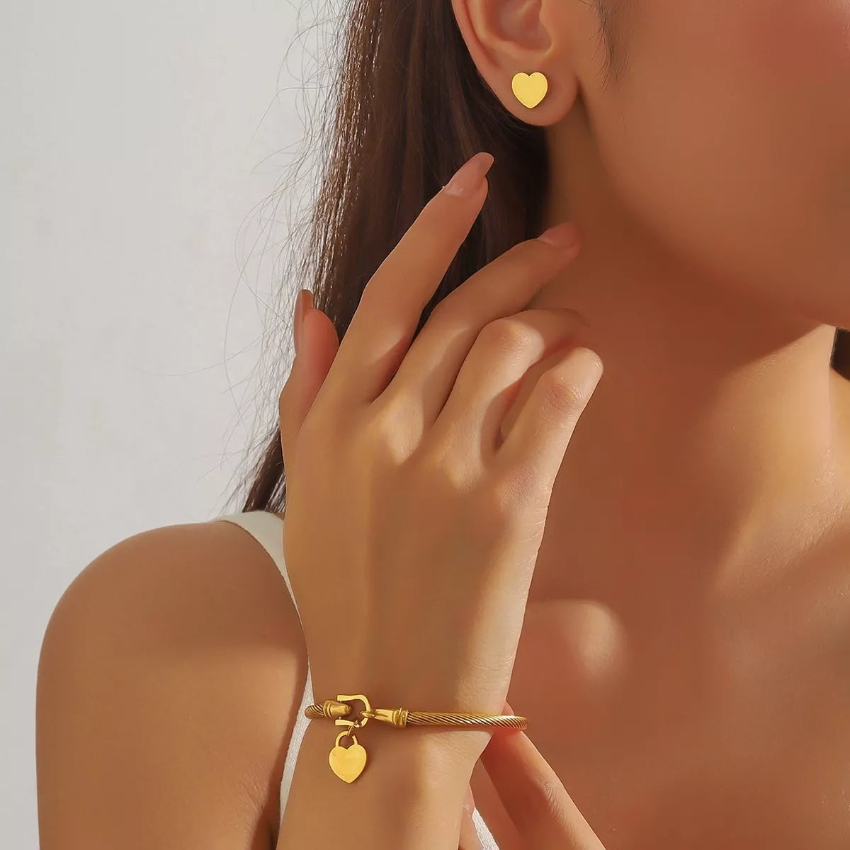 Close-up of a person wearing gold heart-shaped stud earrings and gold bangle bracelet with a heart charm against a neutral background