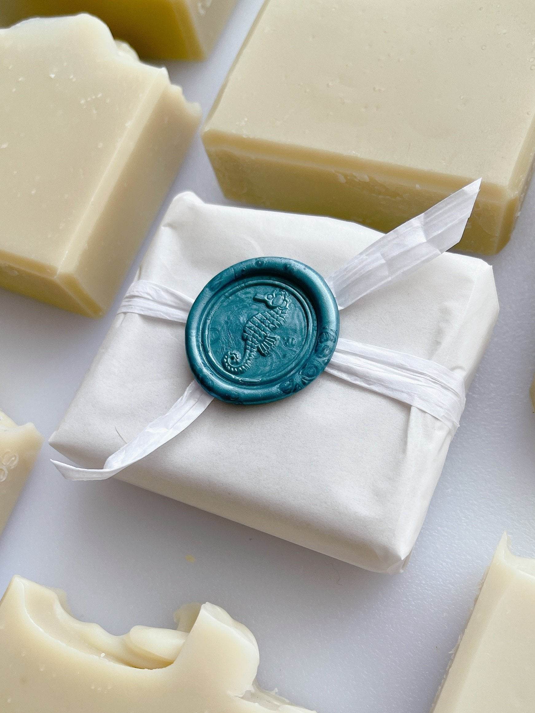 Natural bars of Artisanal Aleppo Style Soap surrounding a soap bar wrapped in white paper with decorative ribbon and blue wax seal on white surface