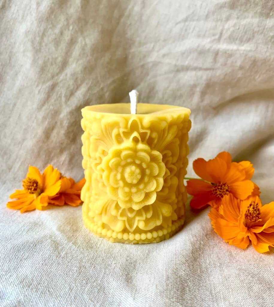 Yellow beeswax pillar candle with flower mandala textured design and orange flowers on light fabric background