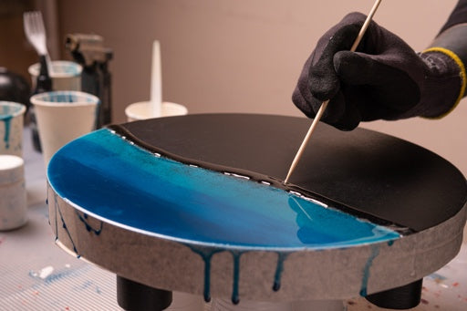 Gloved hand applying blue epoxy resin to round table top with stick in art studio
