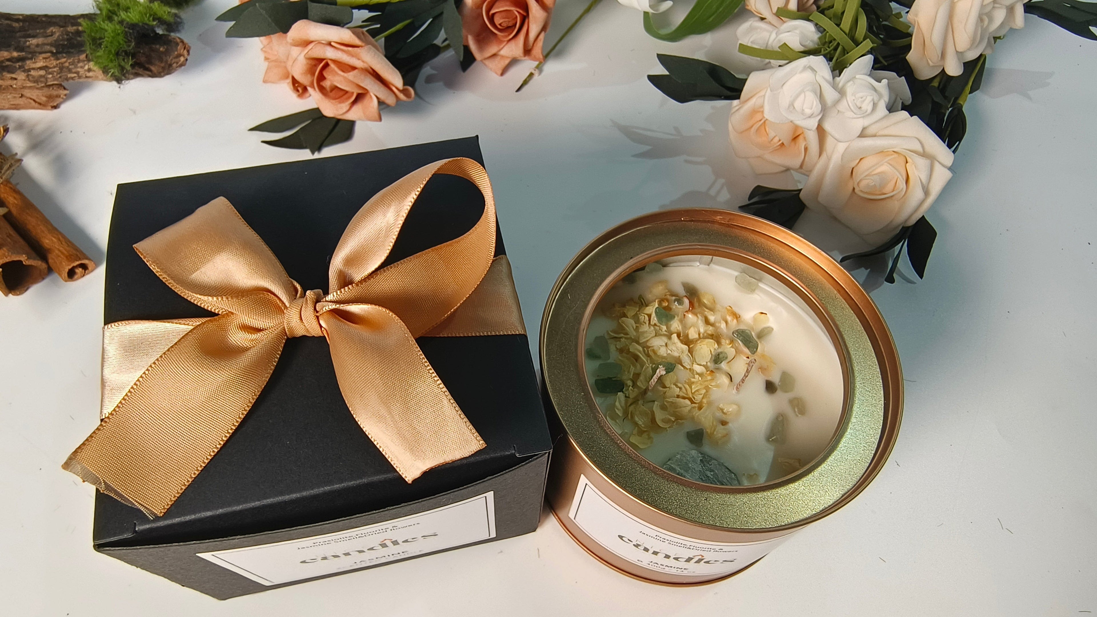 Natural essential oils candle in gold container with black gift box featuring gold bow on white surface with flowers - top view