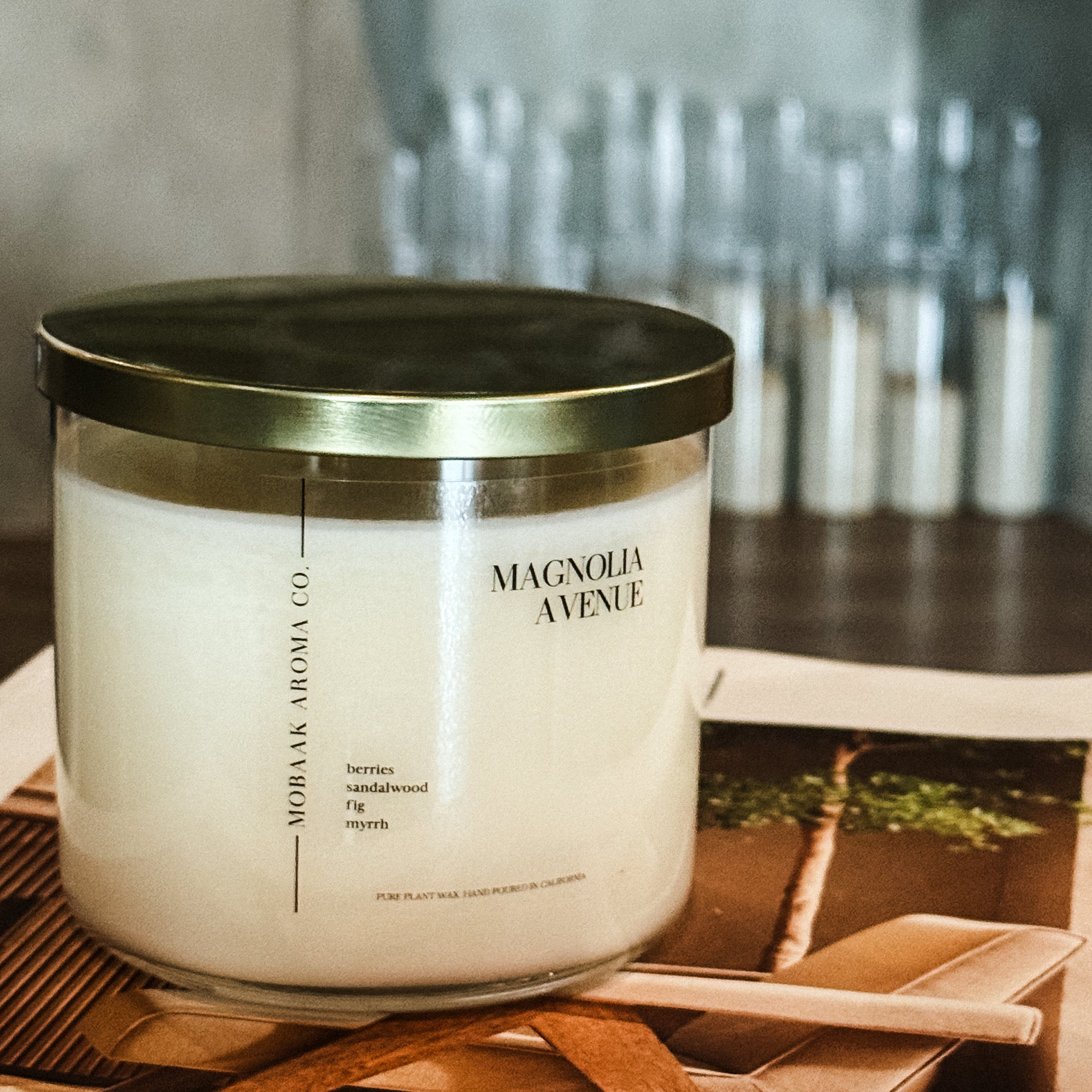Magnolia Avenue soy candle in clear glass jar with gold lid on wood surface - closeup partial front and side view with Mobaak Aromatherapy Shoppe label