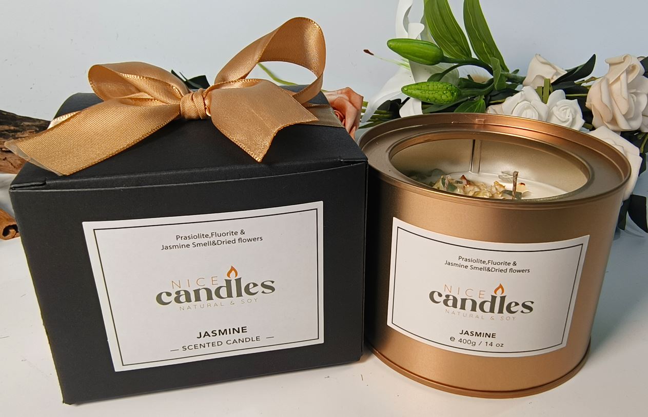 Natural essential oils candle in gold metal container next to black gift box with gold bow labeled jasmine with flowers in background - front view