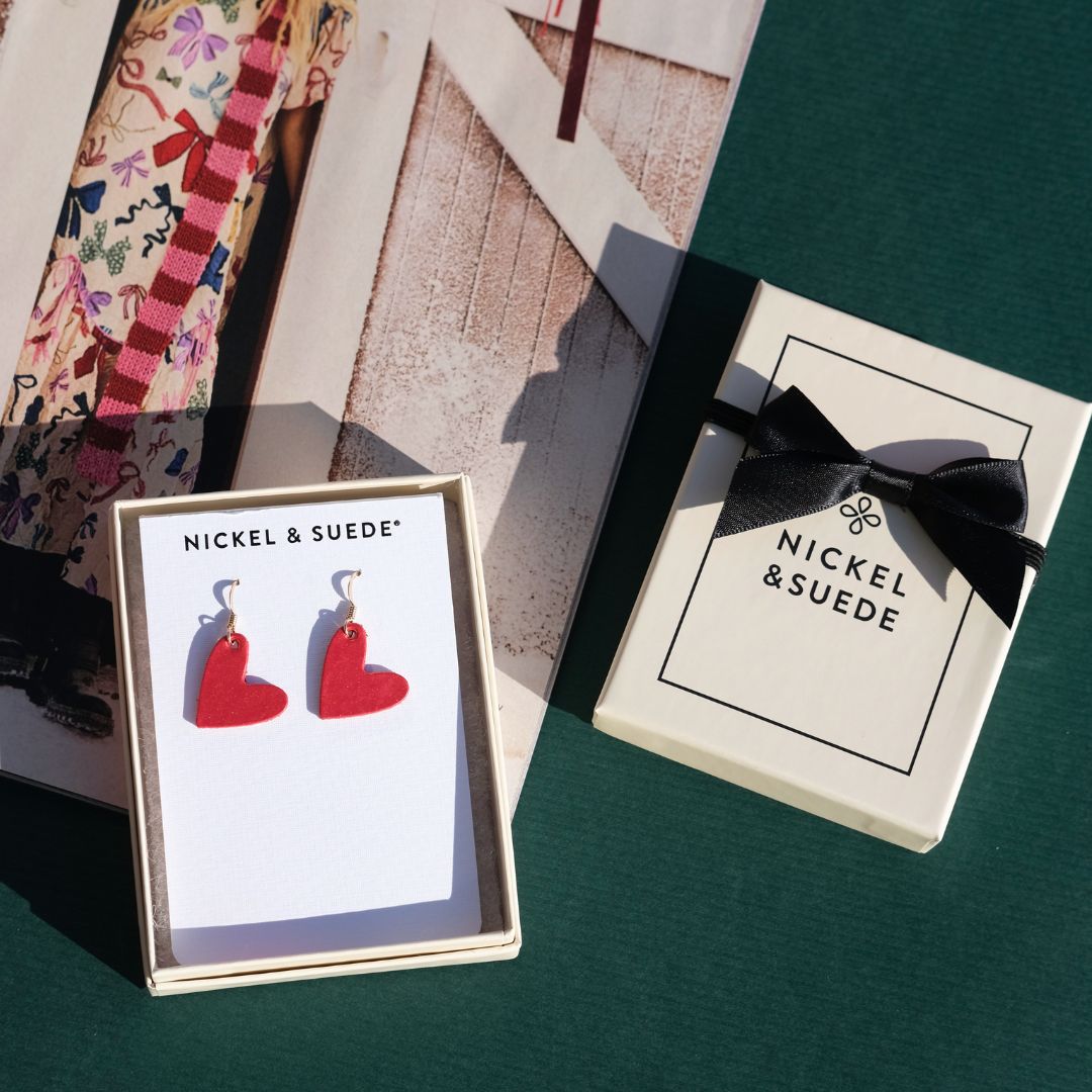 Red heart-shaped earrings in a 'Nickel & Suede' branded box on a green surface