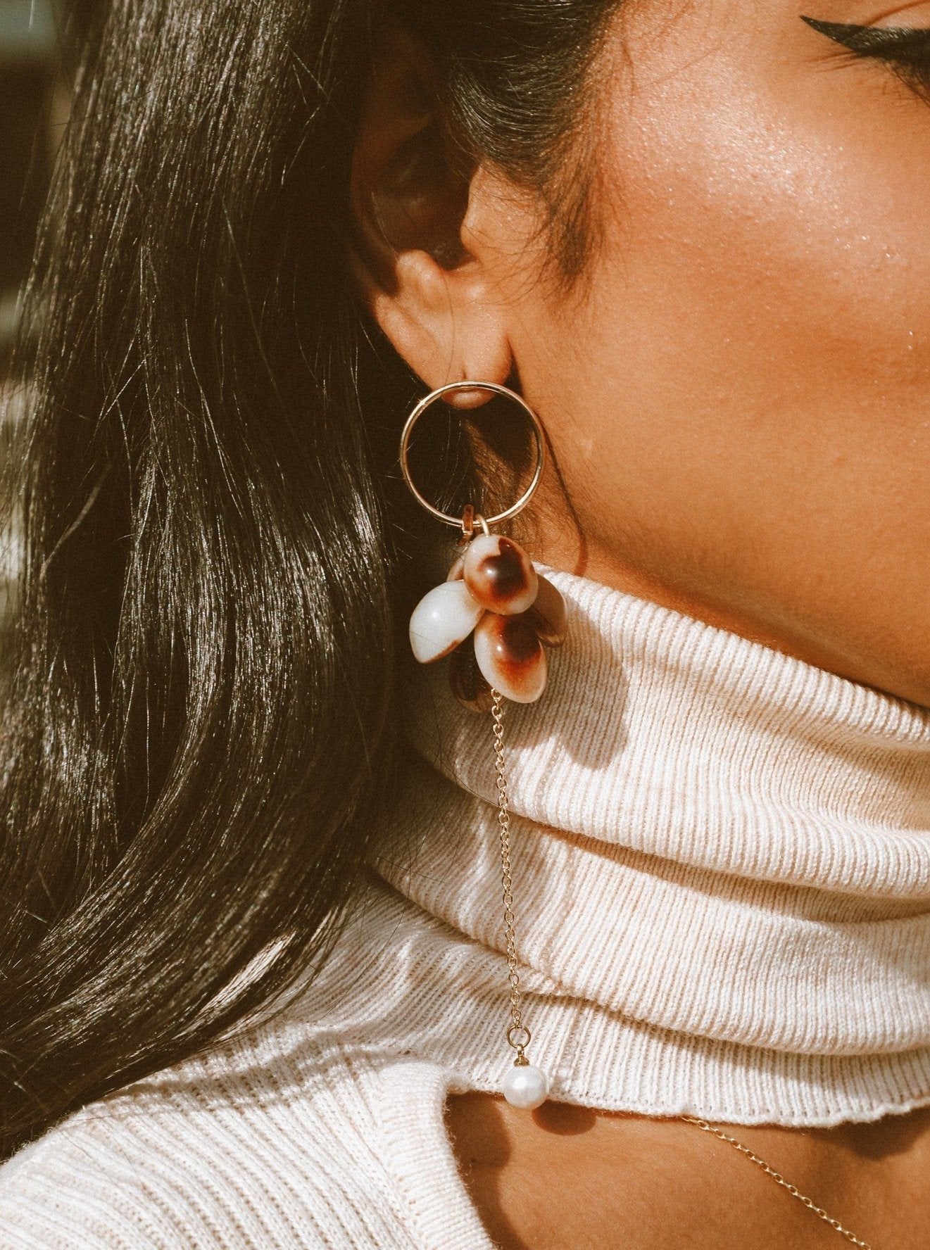 Close-up of a person wearing hoop earrings with dangling shell cluster and pearl on a gold chain against her white turtleneck sweater
